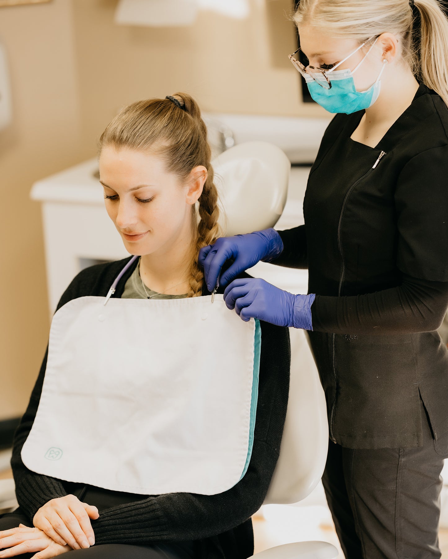 A young woman wearing a mask and gloves fastens a reusable dental bib around another young woman's neck.