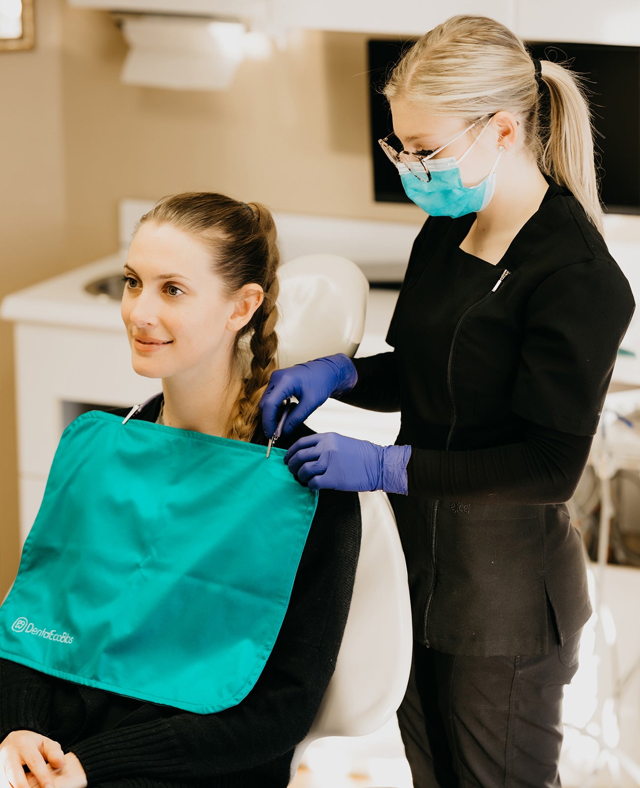 A young woman wearing a mask and gloves fastens a reusable dental bib around another young woman's neck.