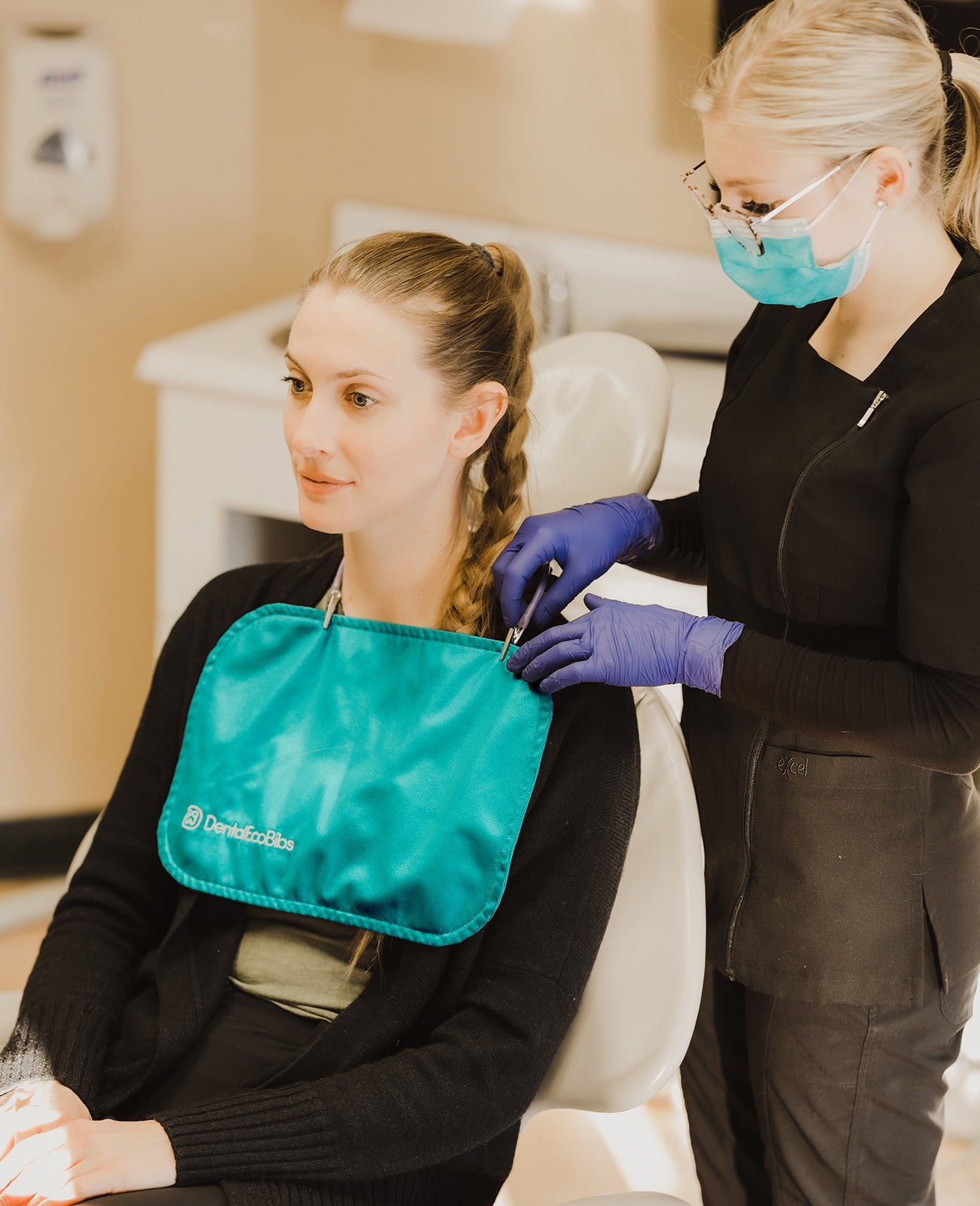 A young woman wearing a mask and gloves fastens a reusable dental bib around another young woman's neck.